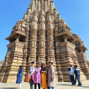 A group of four women smiling in front of the intricately carved Kandariya Mahadeva Temple at Khajuraho under a clear blue sky.