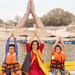 Varanasi women group tour - A woman and two children in life jackets on a boat mimicking the giant Namaste hand sculpture in the background at Prayagraj.