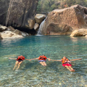 Meghalaya women only group tour - Three people in orange life jackets floating on their backs in a clear turquoise river near a waterfall and large rocks.