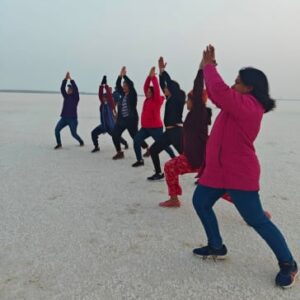 Kutch women group tour with F5 Escapes. A group of women in winter wear performing a yoga pose on the white salt flats of the Rann of Kutch under a pale sky.