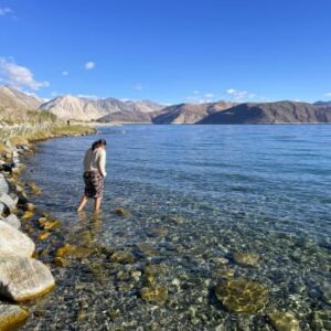 A woman walking in the crystal clear turquoise waters of Pangong Lake with mountain ranges in the background under a blue sky.