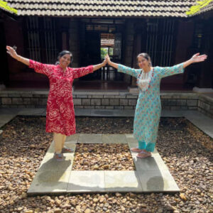 Two women in colorful traditional Indian kurtas posing with outstretched arms in a stone-paved courtyard.