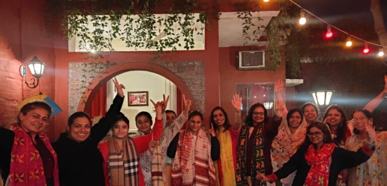 A group of women in colorful festive attire posing together and smiling in front of a red building decorated with string lights at night.