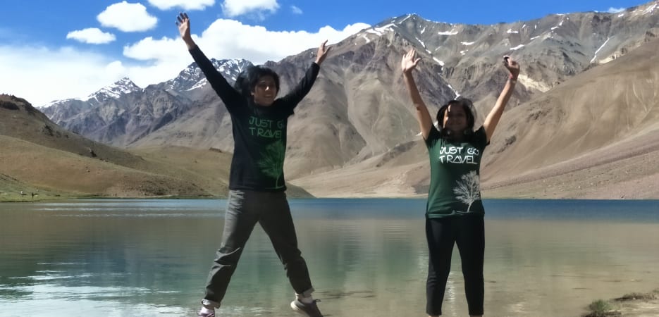 Two women jumping in the air with arms raised beside a serene high-altitude lake surrounded by rugged mountains.