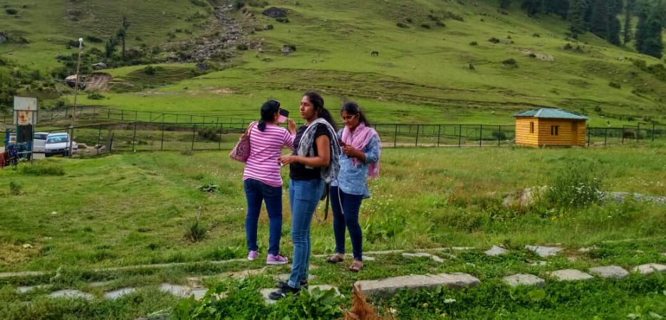 Three young women standing in a lush green meadow with pine forests and mountains in the background.