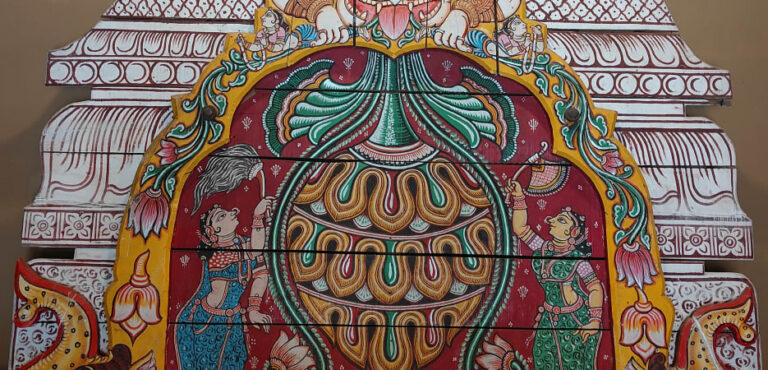 A vibrantly painted wooden temple panel featuring a fierce Kirtimukha deity face, an ornate vessel, and female attendants.