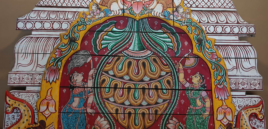 A vibrantly painted wooden temple panel featuring a fierce Kirtimukha deity face, an ornate vessel, and female attendants.