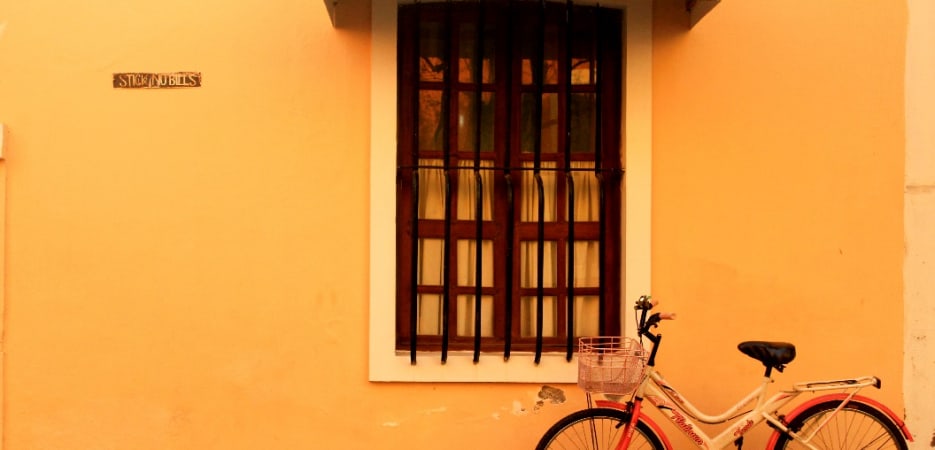 A white and red bicycle with a basket stands in front of a yellow building wall with a barred window.
