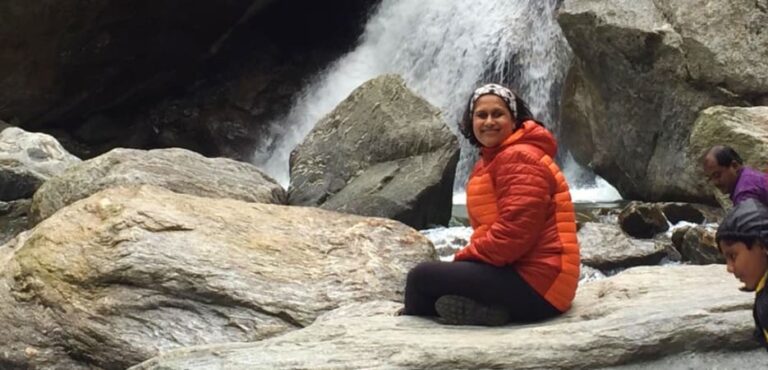 A smiling woman in an orange puffer jacket sits cross-legged on a rock in front of a cascading waterfall.
