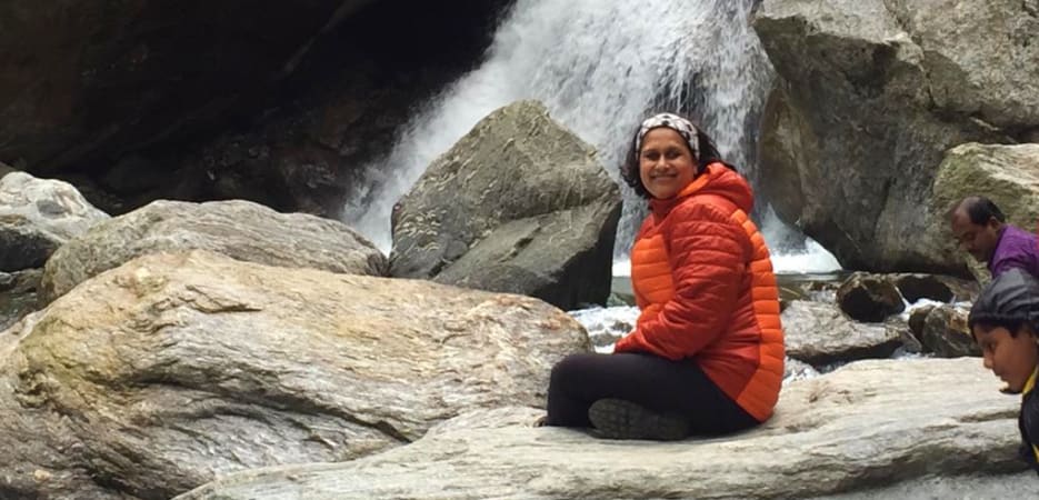 A smiling woman in an orange puffer jacket sits cross-legged on a rock in front of a cascading waterfall.