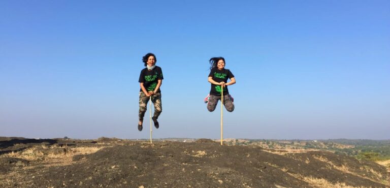 Tips to enjoy group travel - Two women in matching 'Just Go Travel' shirts and camo pants jumping in the air on a rocky hill, holding sticks to create a fun illusion.