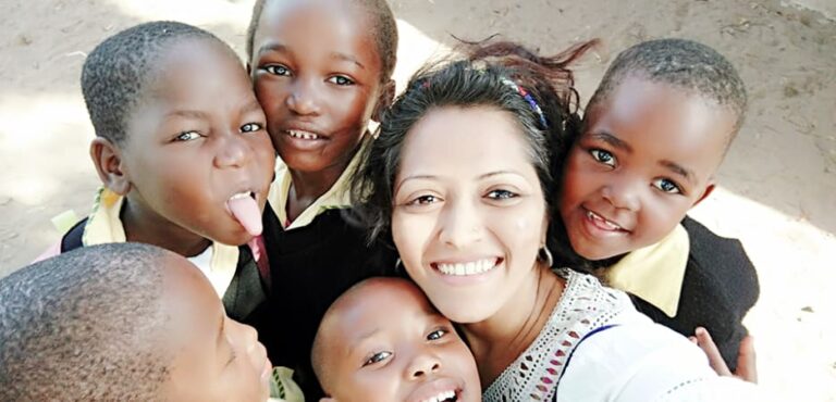 A smiling woman of South Asian descent takes a selfie surrounded by five playful African children in school uniforms.