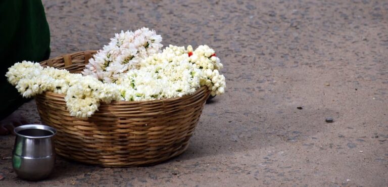 A woven wicker basket overflowing with fresh white jasmine flower garlands sits on a paved street next to a small metal cup.