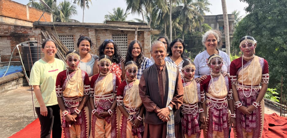 Ethos of F5 Escapes - A group portrait of young Odissi dancers in traditional costumes and jewelry, standing with their teachers and an elder on a red stage outdoors under palm trees.