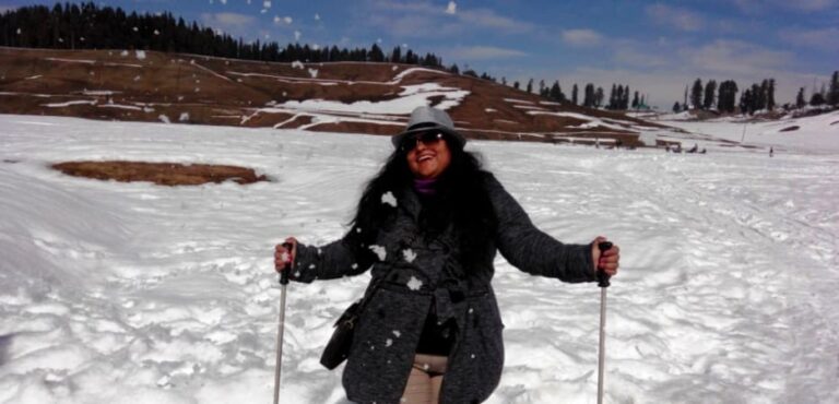 F5 Talks with Pradnya Kulkarni - A smiling woman in a grey coat and hat holds ski poles while snow is tossed in the air around her in a snowy landscape.