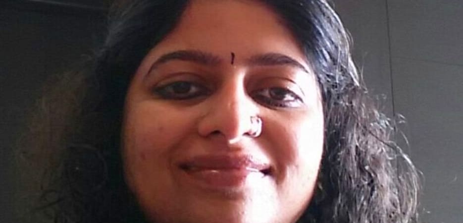 A middle-aged Indian woman with curly hair, a nose ring, and a bindi, smiling at the camera.