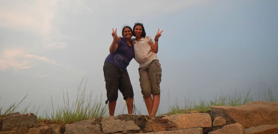 Two smiling women stand together on a stone ledge, making peace signs against a wide, overcast sky.