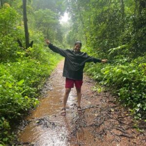 A smiling woman in a black raincoat and red shorts standing on a muddy forest path with arms outstretched.