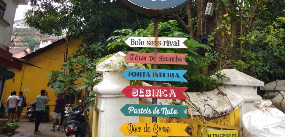 A circular black sign for Confeitaria 31 de Janeiro bakery with directional arrows pointing to various traditional sweets in Fontainhas, Goa.
