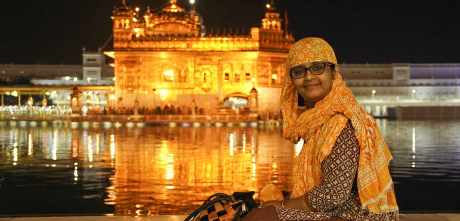 A woman in traditional attire and a headscarf sitting by the Sarovar at the illuminated Golden Temple in Amritsar at night.