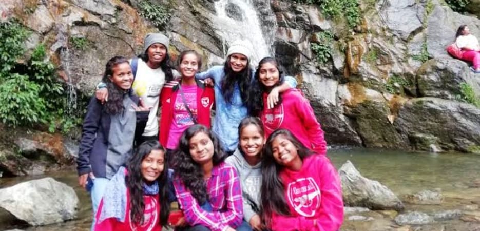 A group of eight young women smiling and posing for a photo in front of a tall waterfall cascading over rocks in a lush forest.