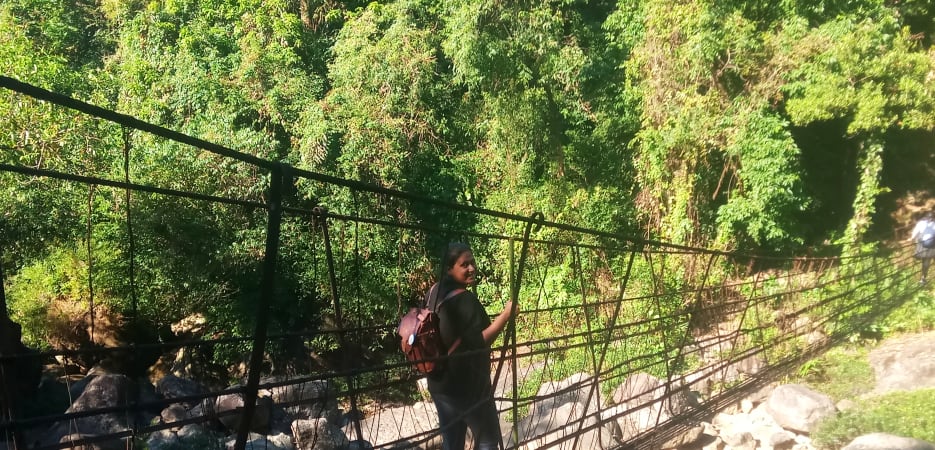 A woman with a backpack walks across a wire suspension bridge over a rocky riverbed in a dense forest.