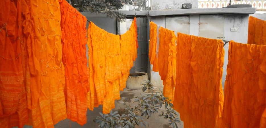 Vibrant orange and yellow tie-dye fabrics hanging on clotheslines to dry in a grey-toned courtyard.