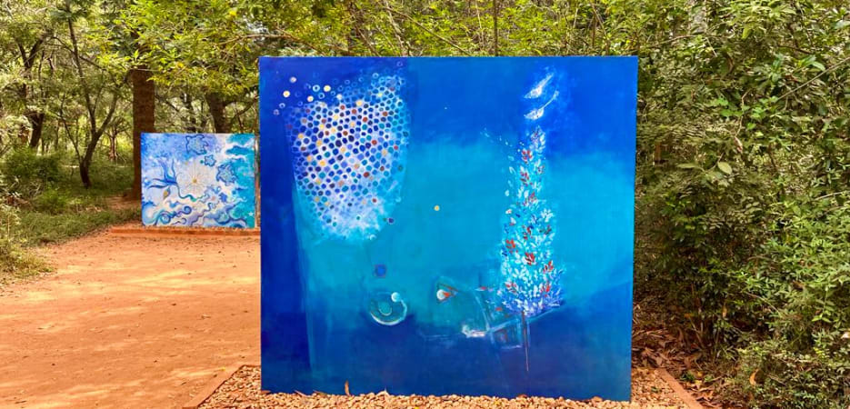 Two large blue abstract art panels standing on a dirt path surrounded by lush green trees in a forest.