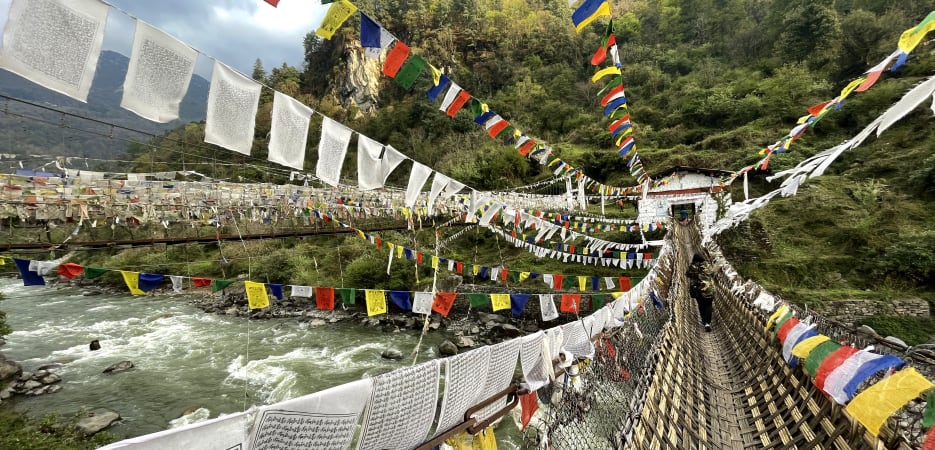 A narrow suspension bridge covered in colorful and white Tibetan prayer flags stretching across a rushing river towards a forested hill.