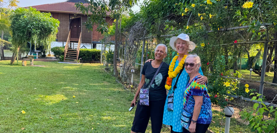 Three smiling women pose together in front of a lush garden and a traditional wooden house on stilts.