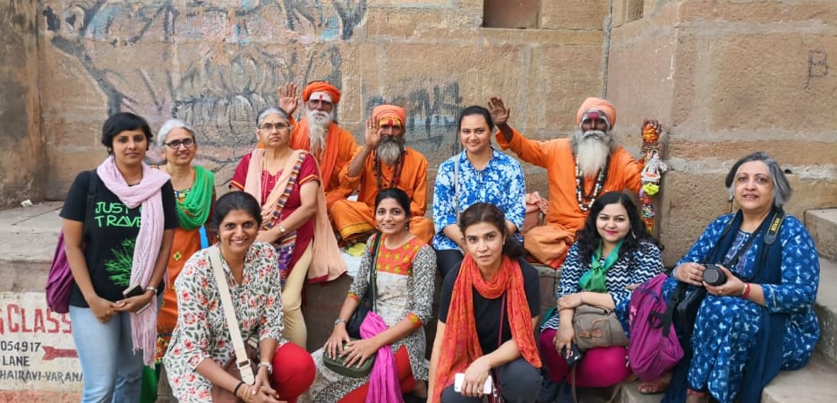 A group of diverse women posing for a photo with three orange-robed sadhus in front of an ancient stone wall in India.
