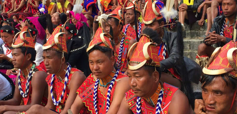 A group of Naga men sitting on stone steps wearing traditional red vests, horned headpieces, and beaded necklaces during a festival in Nagaland.