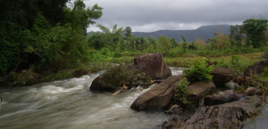 A fast-flowing river cascading over large dark rocks in a lush green tropical forest under a cloudy sky.