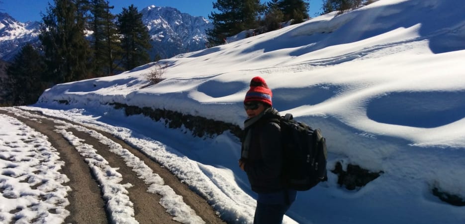 A person with a backpack and winter gear standing on a snow-dusted road in a mountainous region.