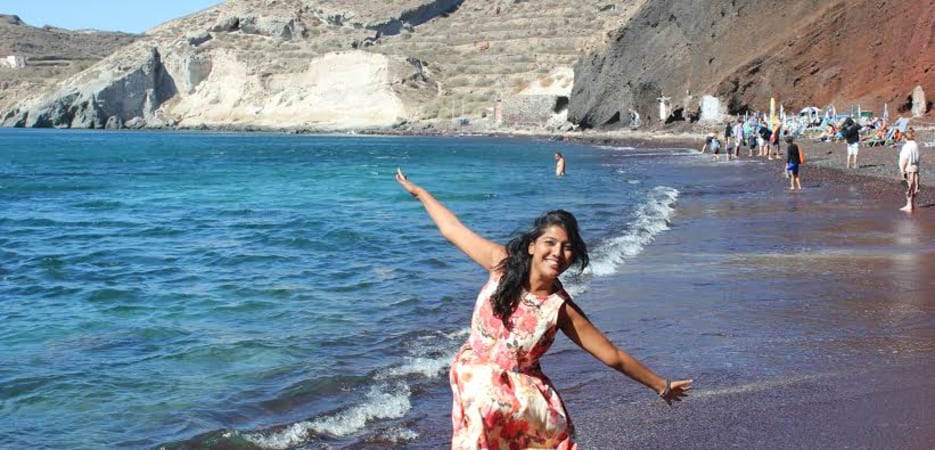 A woman in a floral dress poses happily on the dark sand of Red Beach in Santorini, Greece.