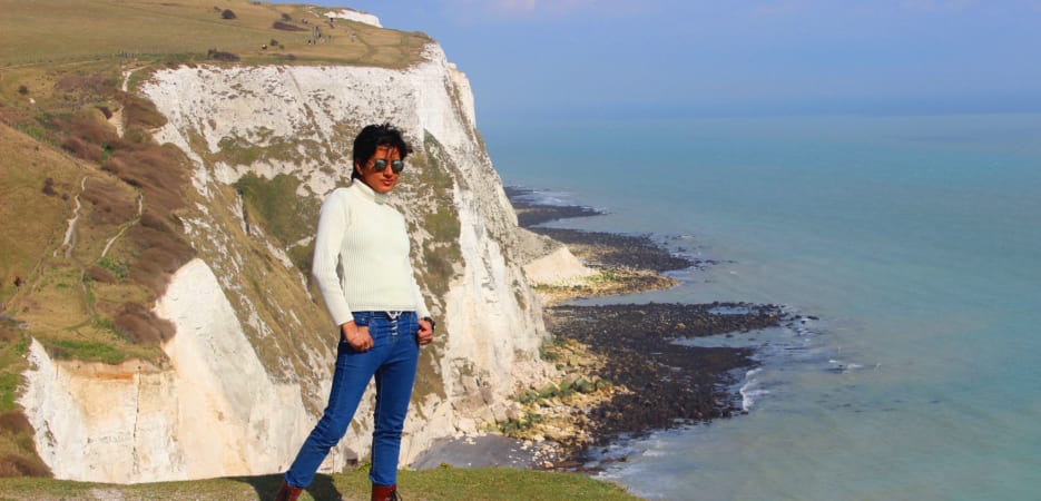 A woman in a white turtleneck and blue jeans standing on a cliff edge at the White Cliffs of Dover with the sea in the background.