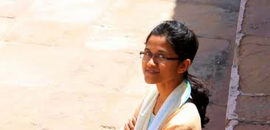 A young woman with glasses and a scarf standing on a stone-paved walkway next to stone steps.