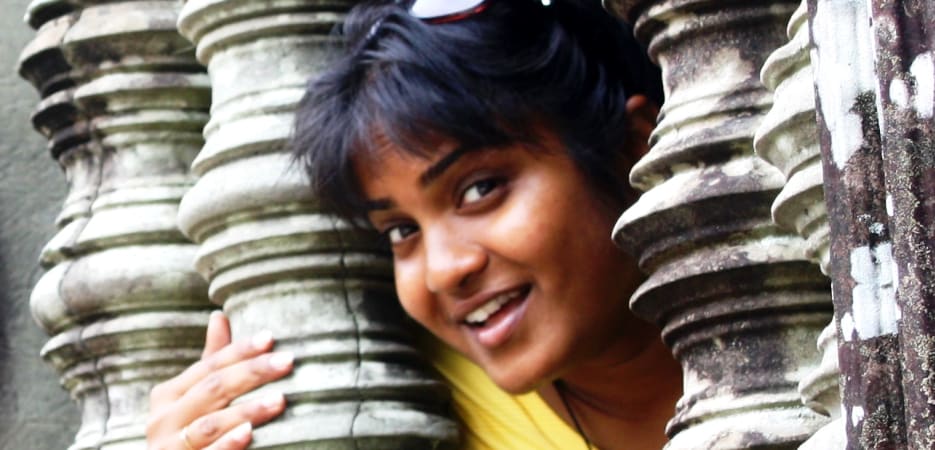 A smiling woman in a yellow shirt peeking through intricately carved stone window bars at an ancient temple.