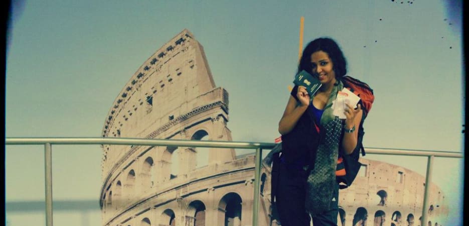 A woman traveler holding her passport and tickets in front of a large mural of the Roman Colosseum.