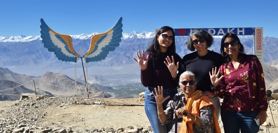 Four women posing and waving in front of a scenic mountain range and an 'I Love Ladakh' sign.