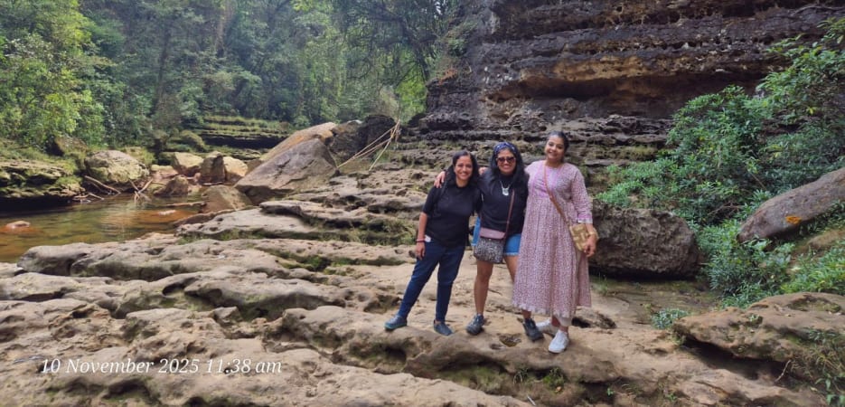Three women smiling together in front of large layered rock walls and a lush forest stream.