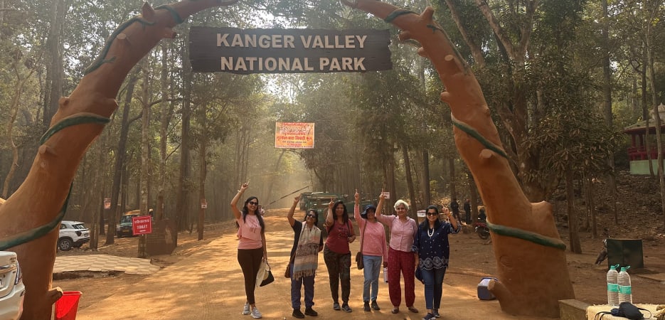 A group of six women pose smiling in front of the rustic wooden entrance sign of Kanger Valley National Park, framed by two large sculpted tree trunks.