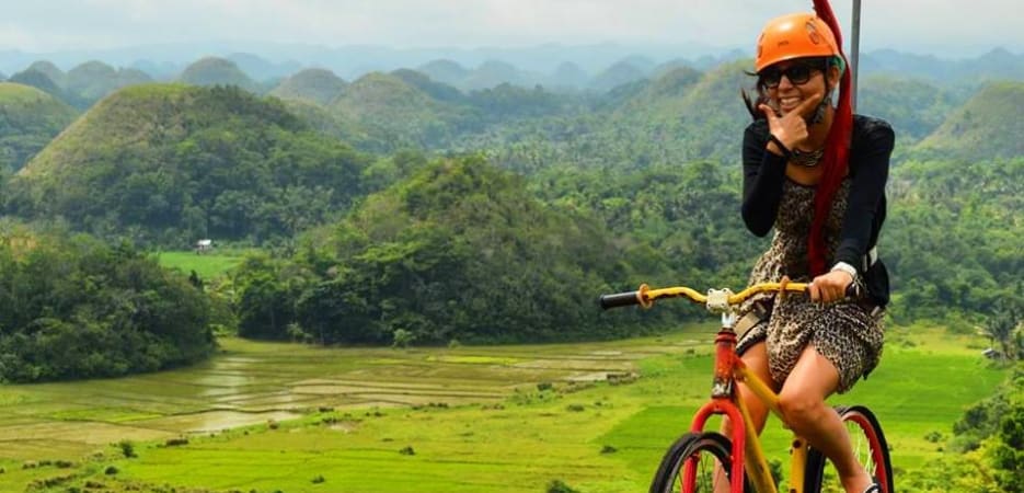 A woman smiles while riding a modified bicycle on a high-wire cable over the Chocolate Hills in Bohol, Philippines.