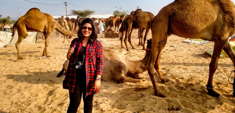 A woman in a red plaid shirt smiling among dromedary camels at a fair in Rajasthan, India.
