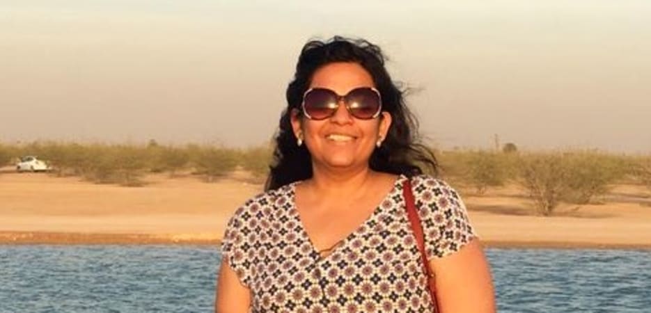 A smiling woman wearing sunglasses and a patterned top stands in front of a body of water in a desert landscape.